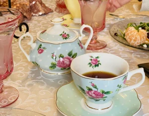 Floral teacup filled with tea on a saucer, beside a matching teapot, set on a white tablecloth. The table also has a salad plate with peeled mandarin slices and pink glassware. A hand holding a pitcher is visible in the background.