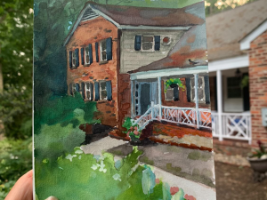 A hand holds a painting depicting a brick house with black shutters, a porch, and a white railing. The house is surrounded by greenery, and a garden is visible in the foreground. The background shows trees and part of another house.