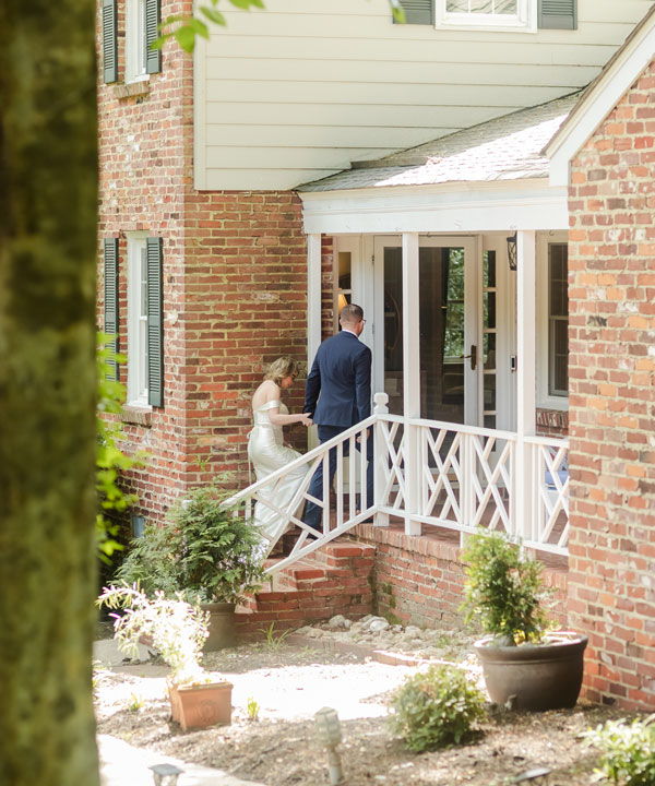 A couple dressed in formal attire walk up the steps to the entrance of a brick Bed and Breakfast in Williamsburg, VA, surrounded by lush plants on a sunny day.